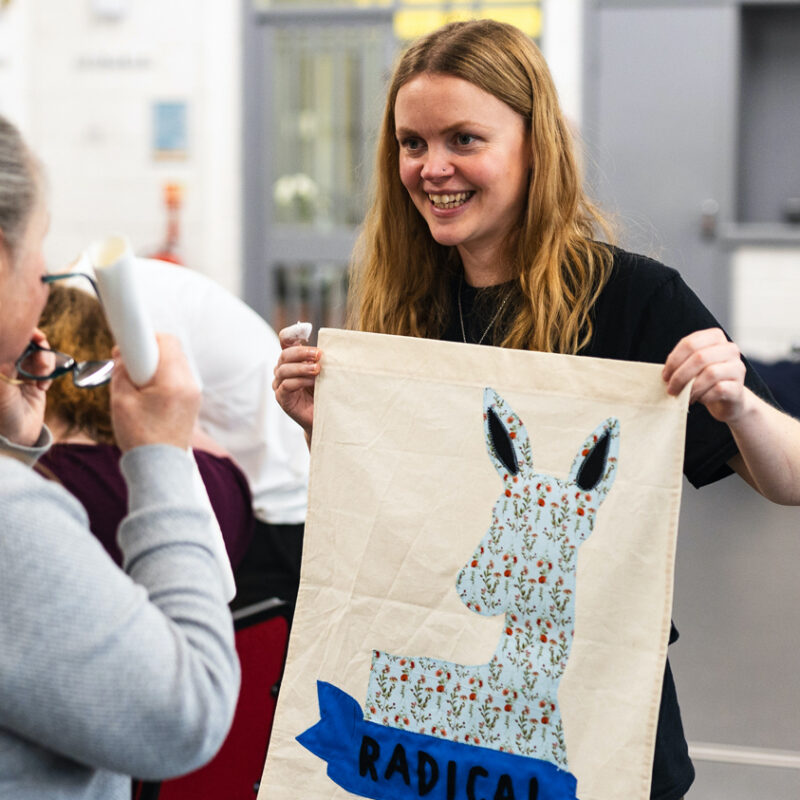 Artist Emily Simpson holds up a small banner with a deer and the words radical on it.