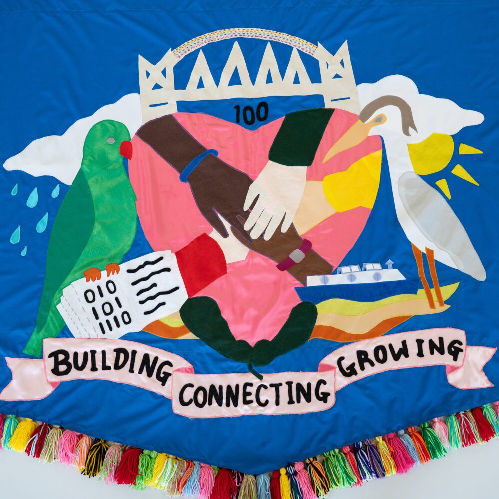 Bright blue banner with multi-coloured tassels, A central bright pink heart with four hands reaching out to one another is embroidered on along with images of wildlife, nature and salford lanmarks,