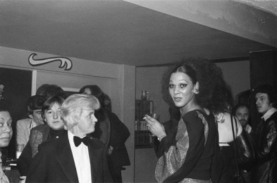 A black and white image by Linda Sterling of a trans woman in a 1970s pub surrounded by a small crowd.