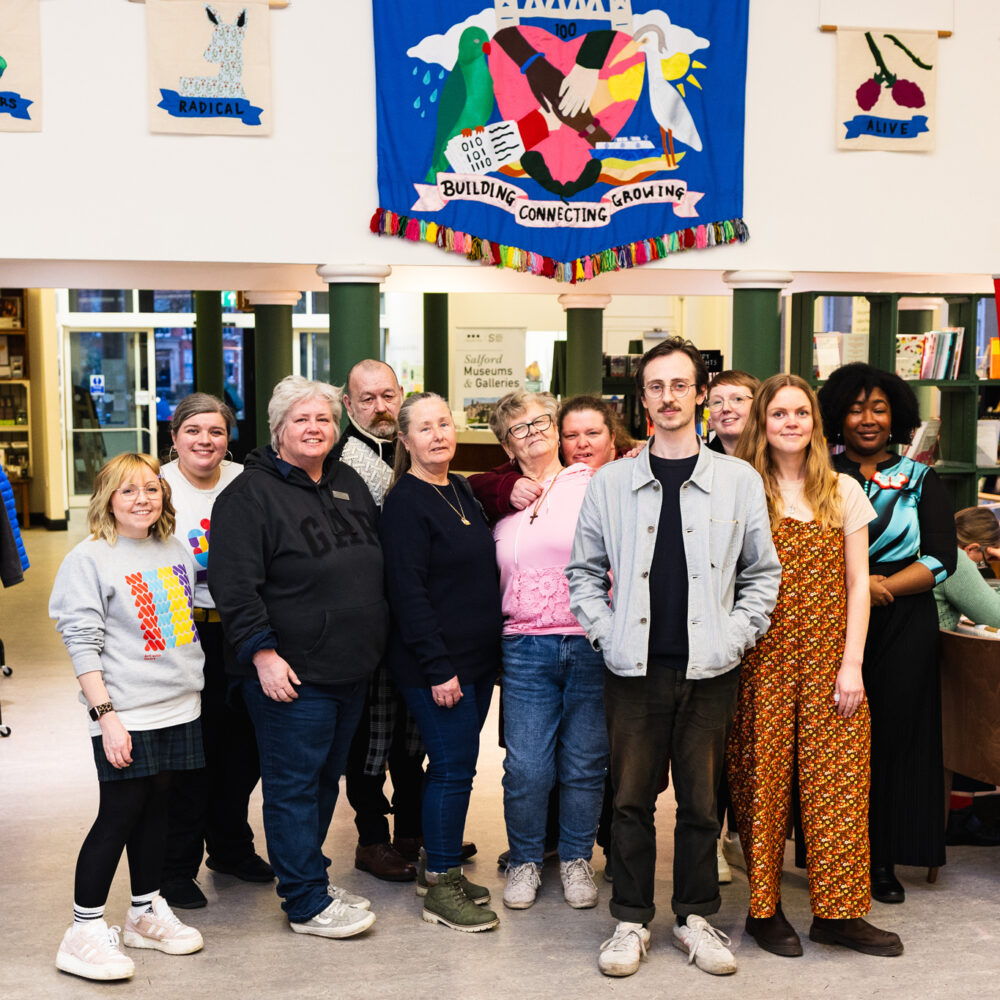 A photo of a group of people at the Salford Voices banner exhibition at Salford Museum and Art Gallery, under a large blue banner.