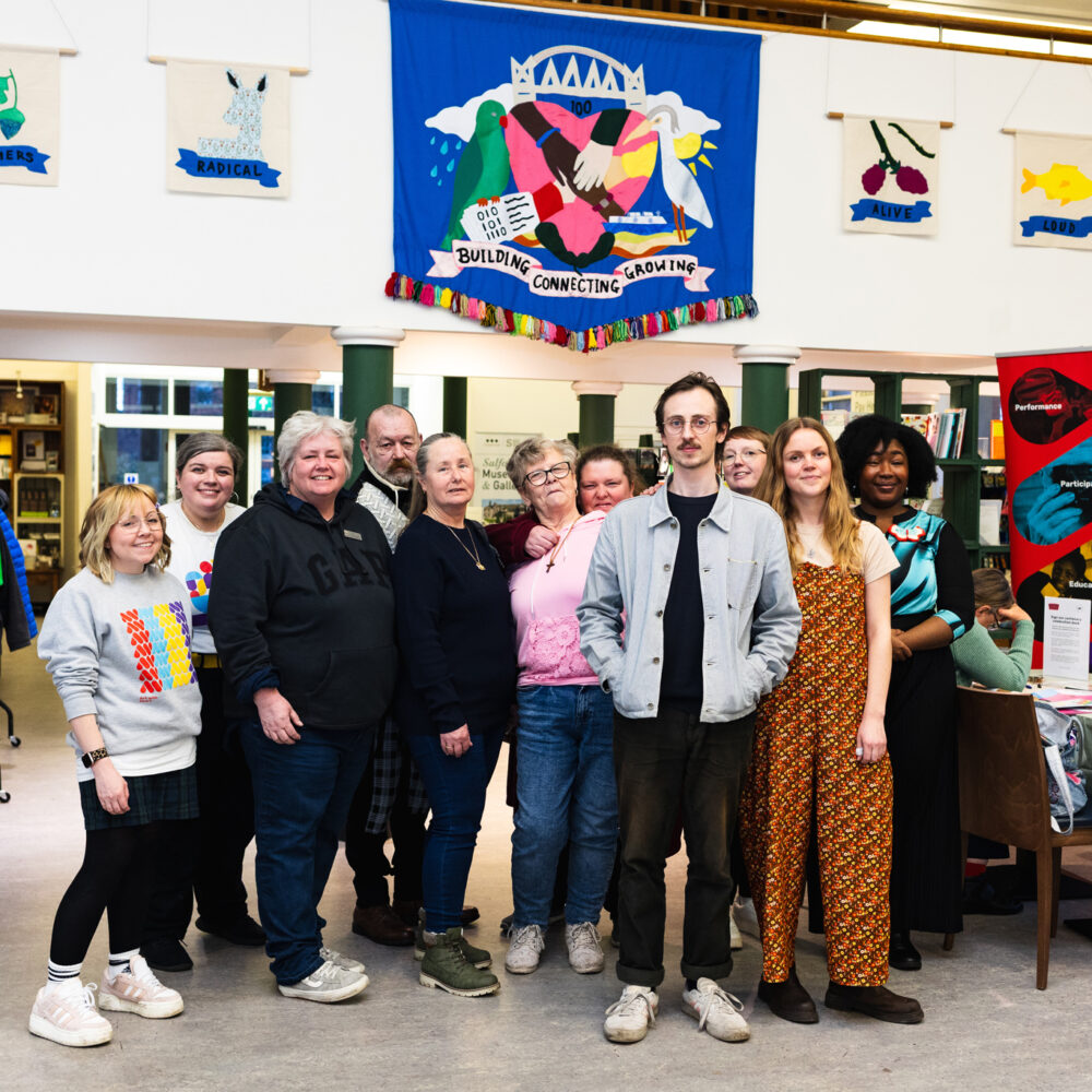 A photo of a group of people at the Salford Voices banner exhibition at Salford Museum and Art Gallery, under a large blue banner.