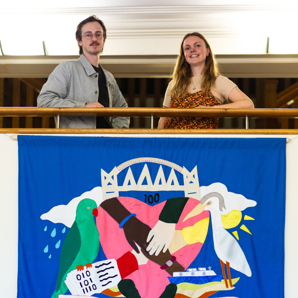 A photo of artists Chris Alton and Emily Simpson at the Salford Voices banner exhibition, stood on a balcony with a big blue banner beneath them that has the words Building, Connecting, Growing on it.