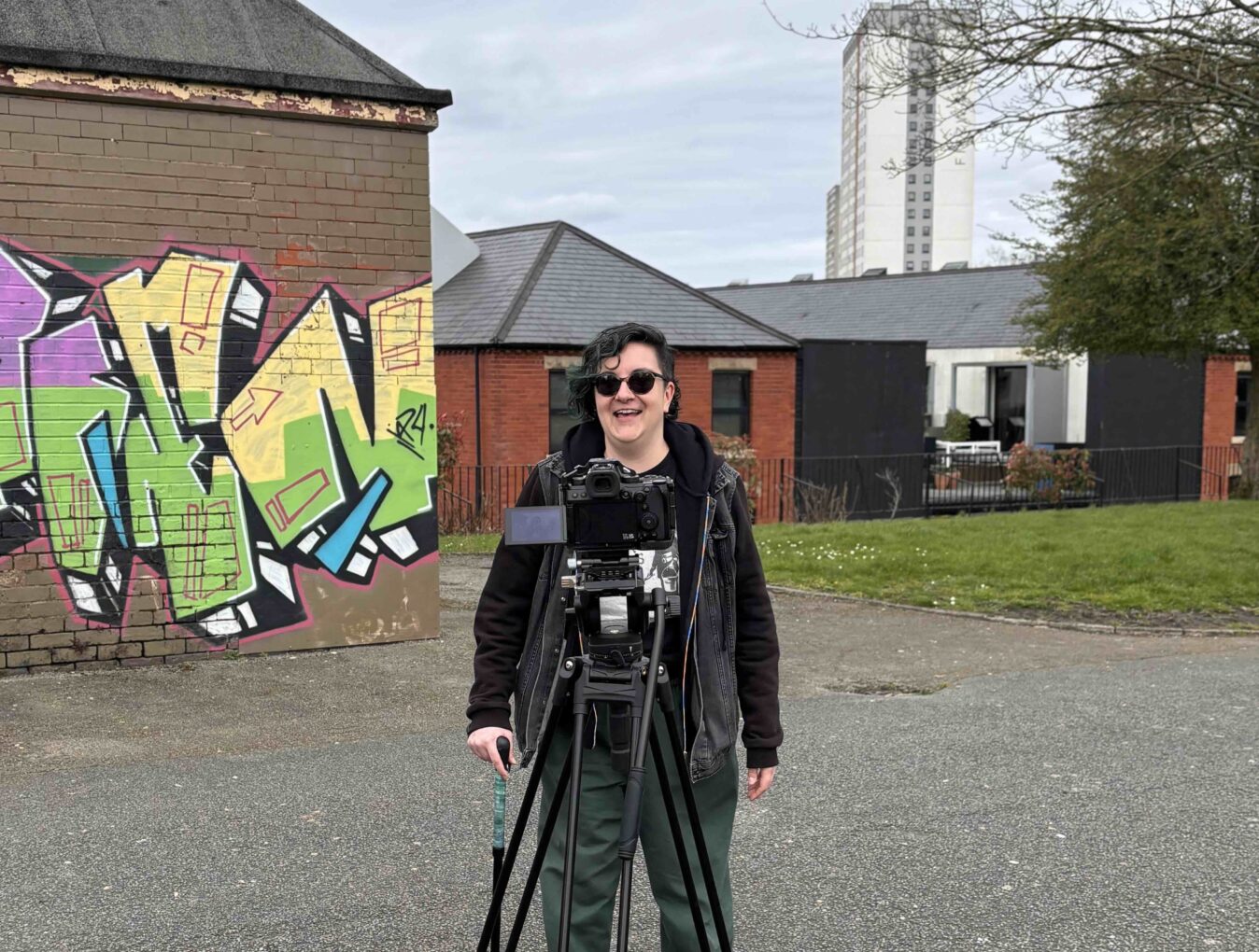 Ash, a Salford born artist stands smiling in front of a film camera in Langworthy park. 