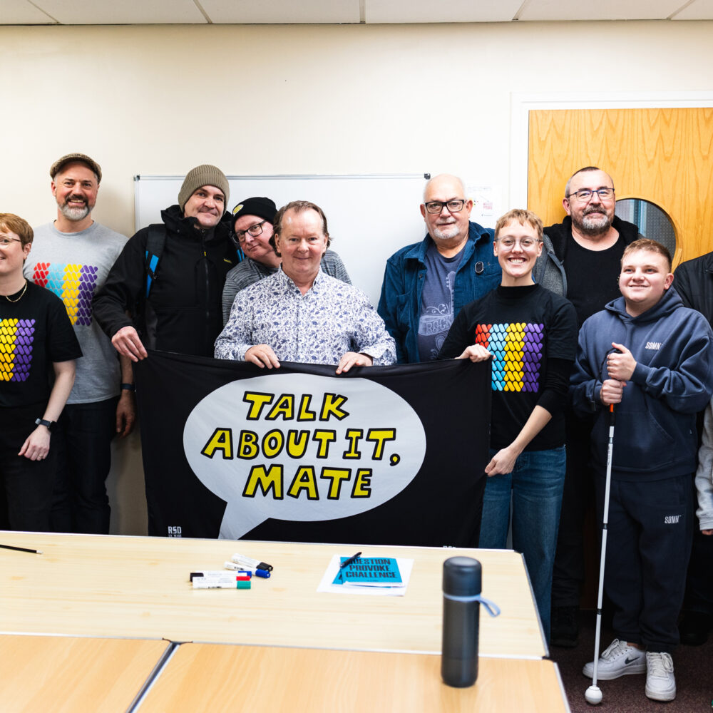A photo of the Talk About It Mate mens mental health group with the Art with Heart team, holding up a banner that says 'Talk About it Mate'.