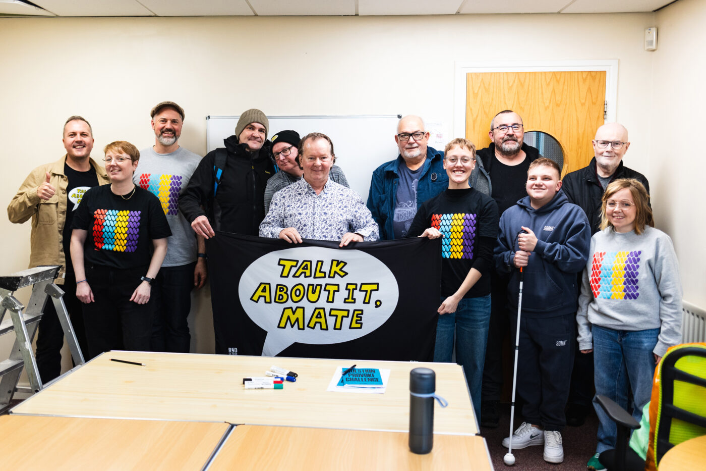 A photo of the Talk About It Mate mens mental health group with the Art with Heart team, holding up a banner that says 'Talk About it Mate'.