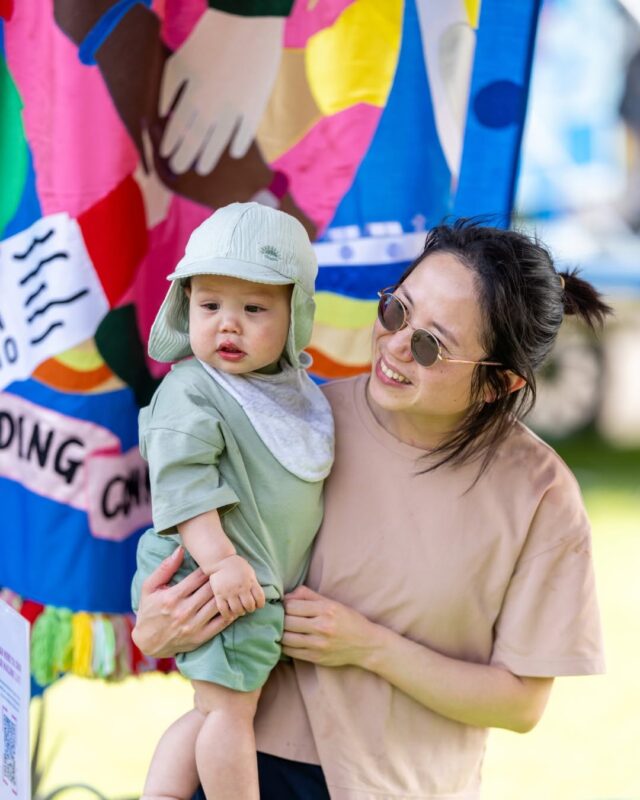 A woman smiles holding a baby in front of our Salford Voices banner