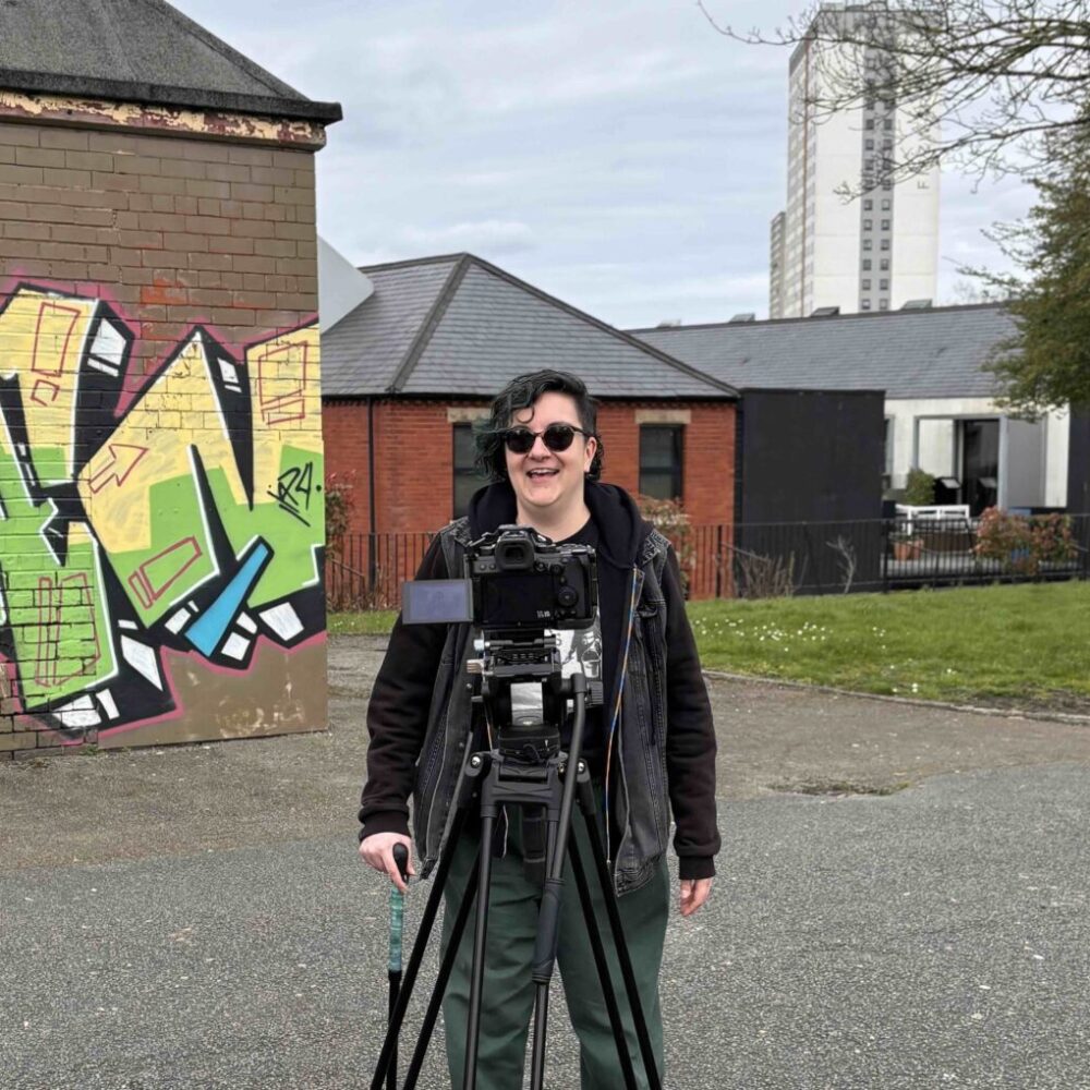 An image of Ash Cox stood behind a camera on a tripod in a yard. There is graffiti on one of the buildings.