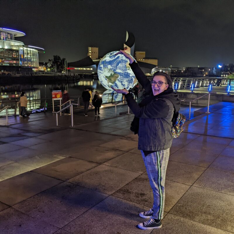 An image of Ash Cox stood in Salford Quays at night, with their arms placed around a large globe in the background.