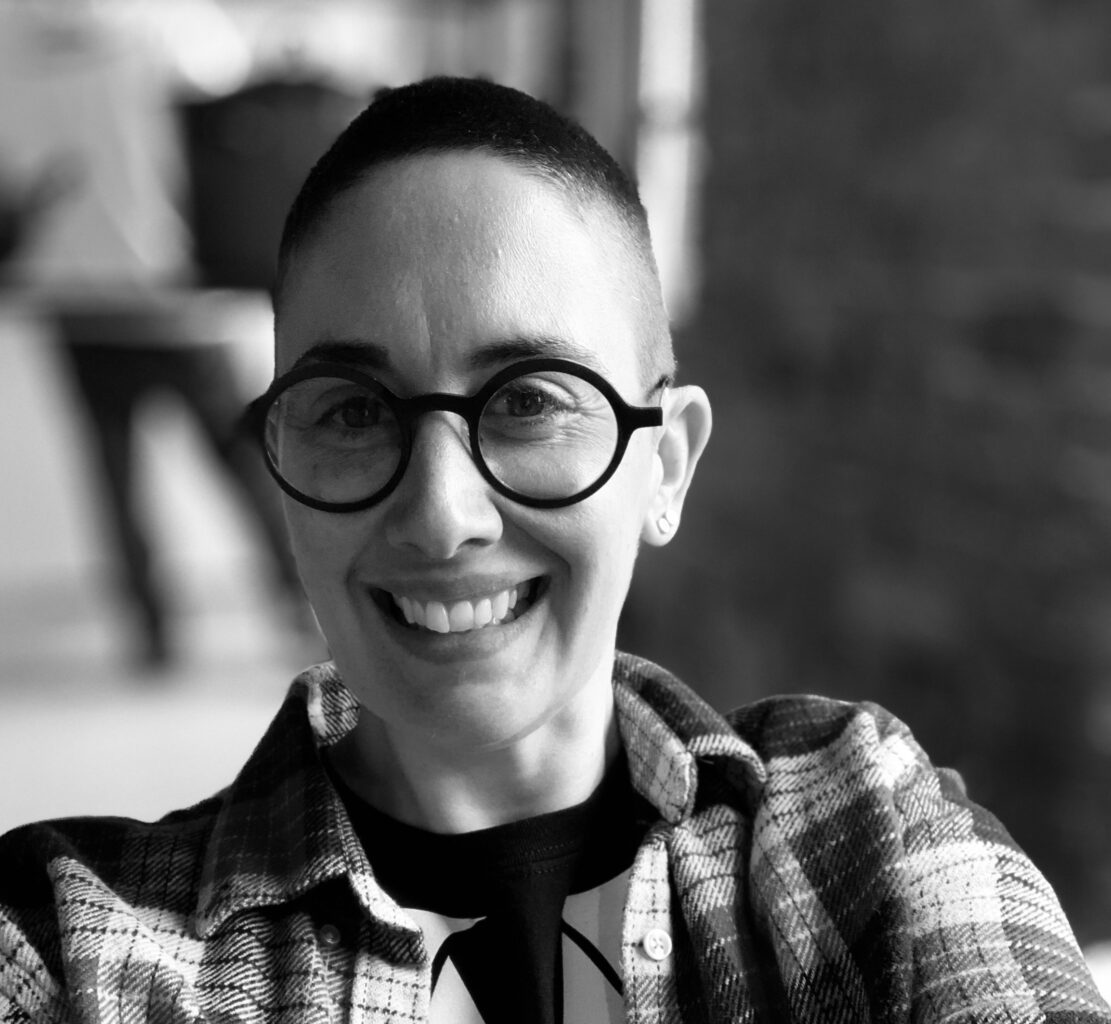 A smiling person with dark hair, olive skin and round black glasses wearing a plaid shirt over a black t-shirt with a necktie printed on it