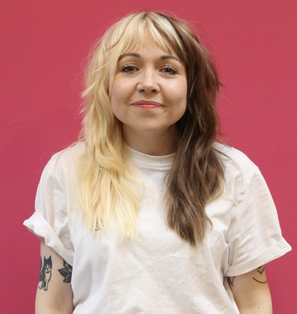 An image of Rose, who has blonde and brown hair and is stood against a pink wall.