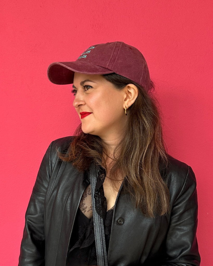 A photo of Natalie Beech, a woman with brown hair stood against a pink wall with a cap on.