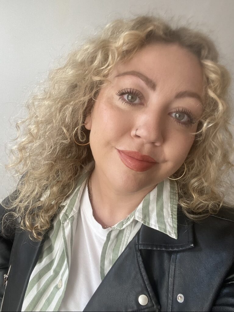 An image of Rebecca, who has long blonde curly hair and is wearing a green and white striped shirt and black leather jacked. She is stood against a white wall.